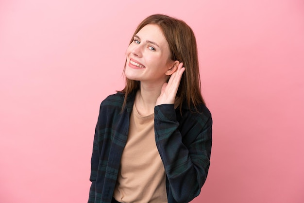Jonge Engelse vrouw geïsoleerd op roze achtergrond die naar iets luistert door hand op het oor te leggen