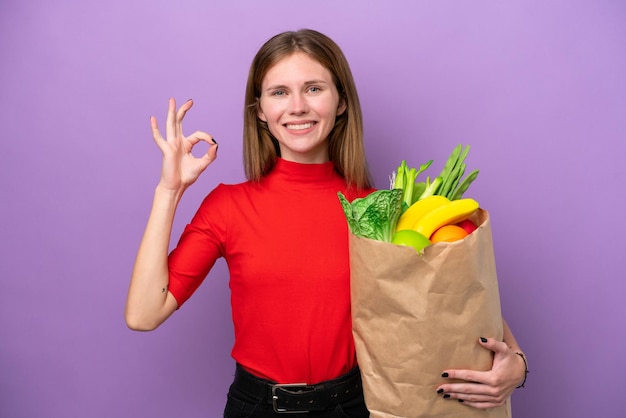 Jonge Engelse vrouw met een boodschappentas geïsoleerd op een paarse achtergrond met ok teken met vingers