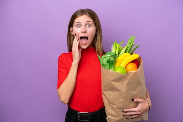 Jonge engelse vrouw met een boodschappentas geïsoleerd op een paarse achtergrond met verrassing en geschokte gezichtsuitdrukking