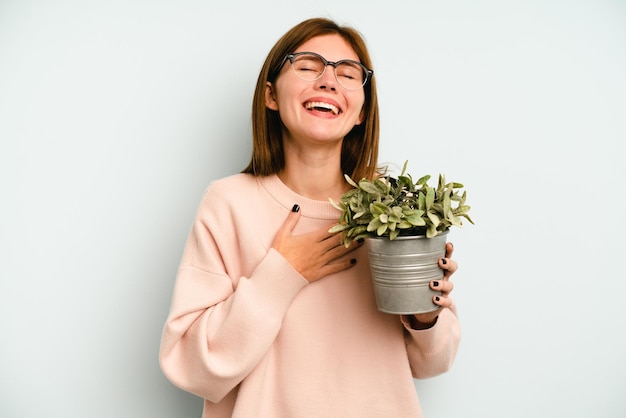 Jonge Engelse vrouw met een plant geïsoleerd op een blauwe achtergrond lacht hardop terwijl ze de hand op de borst houdt.