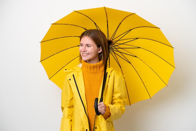 Jonge Engelse vrouw met regenbestendige jas en paraplu geïsoleerd op een witte achtergrond op zoek naar de zijkant en glimlachen