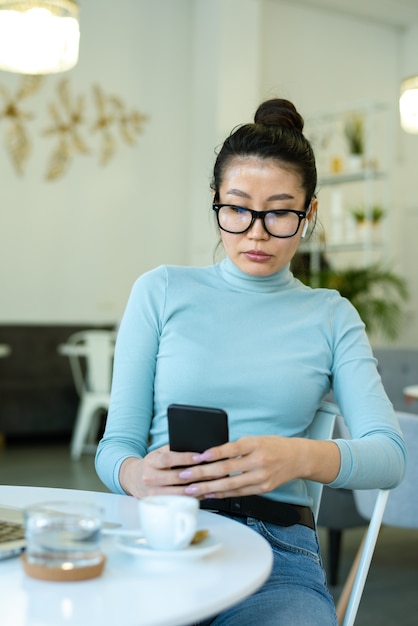 Jonge ernstige vrouw in brillen en vrijetijdskleding scrollen in smartphone terwijl het hebben van koffie of thee aan tafel in café