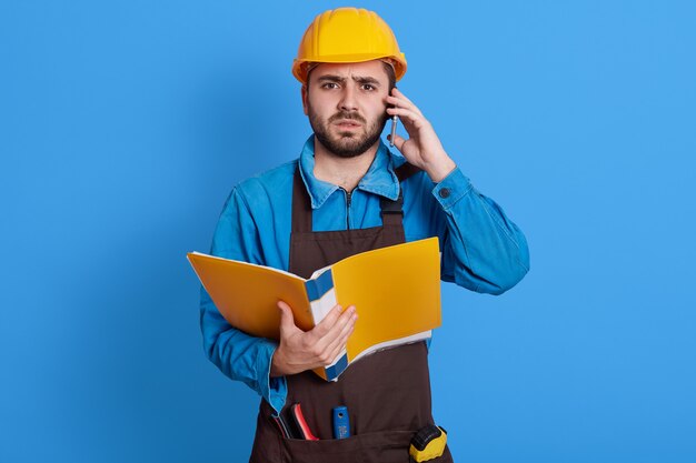 Jonge Europese bebaarde man die als architect of voorman werkt, problemen bij het bouwen oplost, een papieren map vasthoudt, een pratende telefoon, een schort, een helm en een blauw uniform draagt.