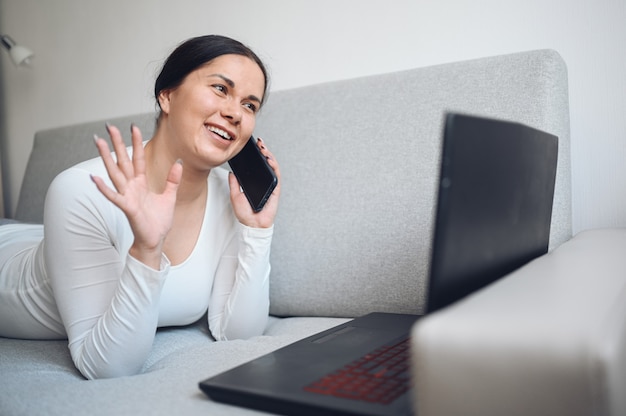 Jonge Europese freelance vrouw die een selfie of videogespreklaptop en telefoon op grijze laag maken tijdens de quarantaine van het coronavirusisolatiehuis. Covid-19 pandemisch Corona-virus. Online werk vanuit huis concept.