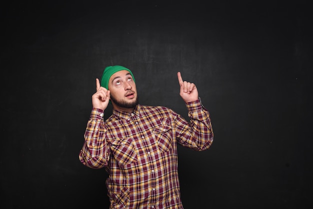 Jonge Europese man met baard in groene gebreide muts, kijkt verbaasd en verbaasd. Vingers naar boven en naar rechts laten zien. Zwarte achtergrond, lege kopie ruimte voor tekst of advertentie