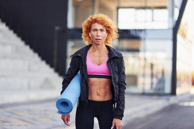 Jonge Europese roodharige vrouw in sportieve kleding met mat in handen buitenshuis