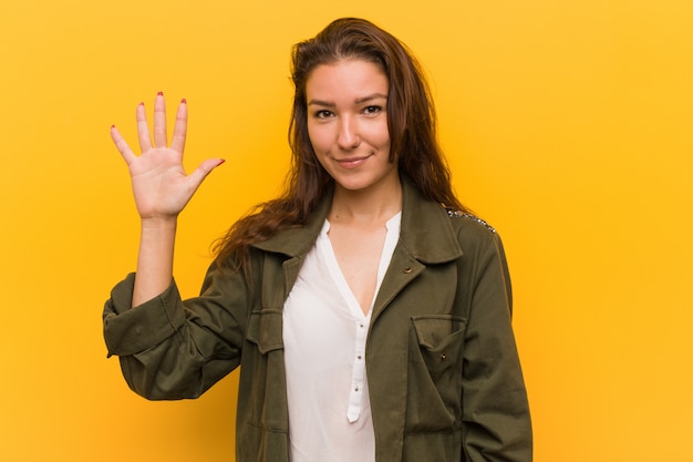 Jonge Europese vrouw die over gele muur wordt geïsoleerd die vrolijk tonend nummer vijf met vingers glimlacht.