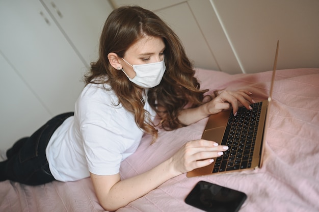 Jonge Europese vrouw in het masker van de gezichtsgeneeskunde die aan laptop en telefoon in bed werken