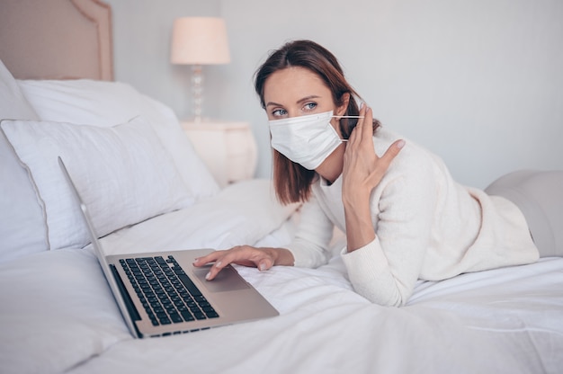 Jonge Europese vrouw in het masker van de gezichtsgeneeskunde die aan laptop in slaapkamer tijdens coronavirusisolatie werken