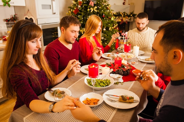 Jonge familie en vrienden bidden en zegenen de tafel tijdens het kerstdiner. Familie viert kerstavond met traditionele gerechten en decoratie thuis