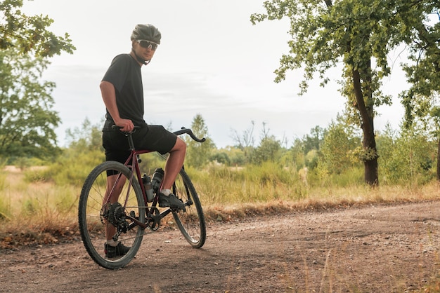 Jonge fietser in bril en helm staat met een fiets op een onverharde weg op het platteland.