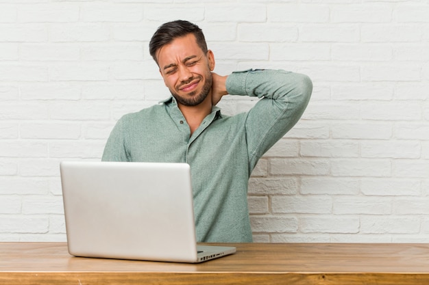 Jonge filipijnse mensenzitting die met zijn laptop werken die aan nekpijn lijden vanwege sedentaire levensstijl.