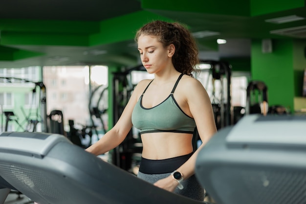 Jonge fit vrouw in sportkleding voordat basistraining draait op een loopband in de sportschool. Het concept van een gezonde levensstijl, warming-up, fitness.