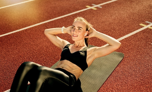 Jonge fit vrouw presteert haar lichaam draaiende oefening voor buikspieren op de mat bij stadion track met rode coating op zonnige dag