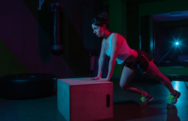 Jonge fit vrouw push-ups uit een houten kist in neon kleurovergang rood blauw licht op een donkere muur Functioneel trainingsconcept.