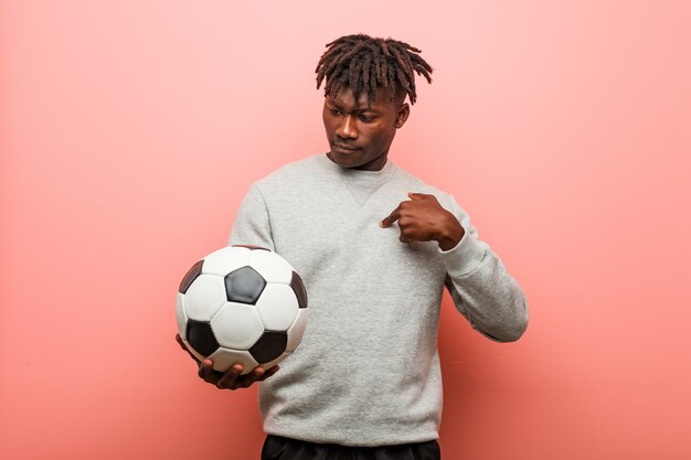 Jonge fitness zwarte man met een voetbal verrast wijzend op zichzelf, breed glimlachend.