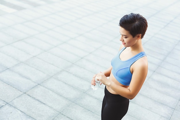 Jonge fitnessvrouw in oortelefoons heeft pauze, drinkt water na training in de stad, kijkt omhoog en glimlacht naar de camera, kopieert ruimte