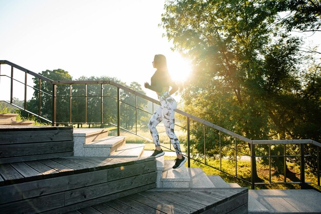 Jonge fitnessvrouw in stijlvolle sportkleding rashguard en leggings die op de trap loopt in een prachtig groen park bij zonsondergang Concept van actieve en gezonde levensstijl