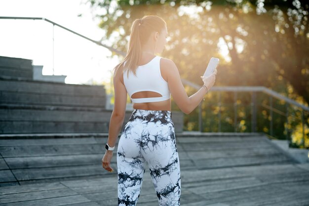 Jonge fitnessvrouw in witte sportkleding die selfie maakt met haar smartphone terwijl ze op een zonnige dag buiten op een houten trap staat Concept van actieve en gezonde levensstijl