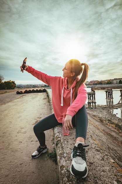Jonge fitnessvrouw rust na een zware training op de muur bij de rivierbrug en neemt selfie met smarthphone.