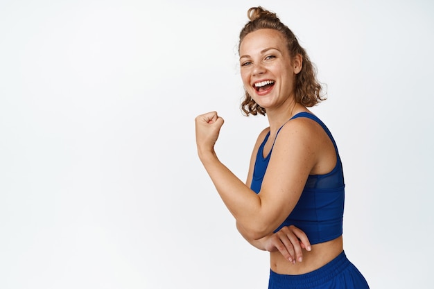 Jonge fitnessvrouw toont spieren, flex biceps en lachen, opscheppen met sterk lichaam na training, sportactiviteiten op wit.