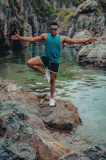 Jonge fitte man met zonnebril die yoga doet door op één been op de rots in de oceaan te balanceren