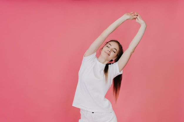 Jonge fitte spectaculaire vrouw strekt zich uit op een roze achtergrond, gekleed in een witte T-shirt witte spijkerbroek