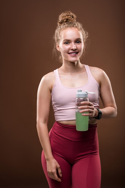 Jonge fitte vrouw in trainingspak met fles met drankje terwijl ze een korte pauze heeft na sporttraining op bruin geïsoleerd