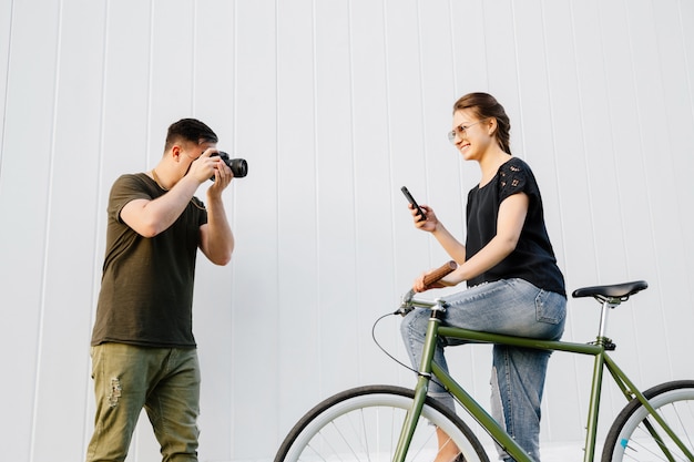 Jonge fotograaf die foto van modieuze mooie vrouw nemen die zich met fiets bevinden en een smartphone gebruiken. Buitenshuis.