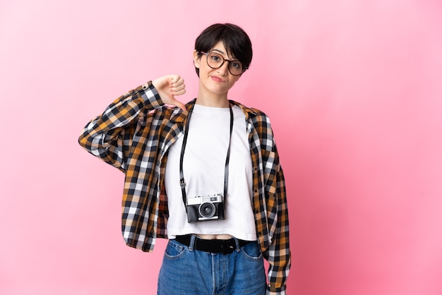 Jonge fotograafvrouw die op roze ruimte wordt geïsoleerd die duim met negatieve uitdrukking toont