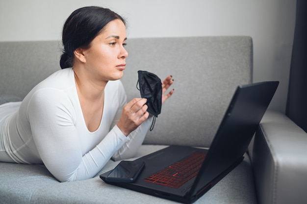 Jonge freelance vrouw die in het masker van de gezichtsgeneeskunde videotelefoneren met laptop op grijze laag tijdens de quarantaine van het coronavirusisolatiehuis. Covid-19 pandemisch Corona-virus. Online werk vanuit huis concept.