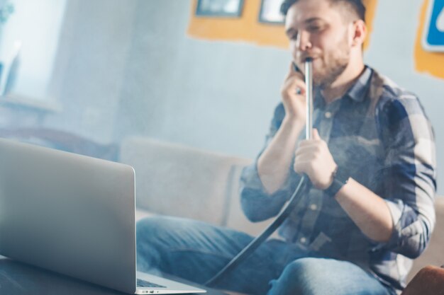 Jonge freelancer die een shisha rookt en op een smartphone spreekt terwijl het werken van huis