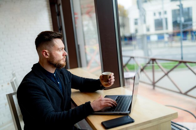 Jonge freelancer of zakenman werkt op laptop en drinkt koffie in een café. Grote panoramische ramen aan de voorkant. Detailopname.