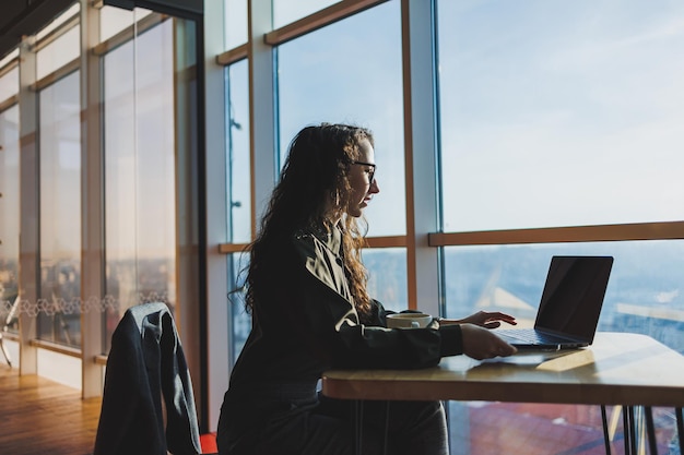 Jonge freelancer vrouw Jonge vrouw die werkt met laptop in coworking ruimte en telefoneert terwijl ze in de open ruimte zit met een kopje koffie en glimlacht terwijl ze een bril vasthoudt