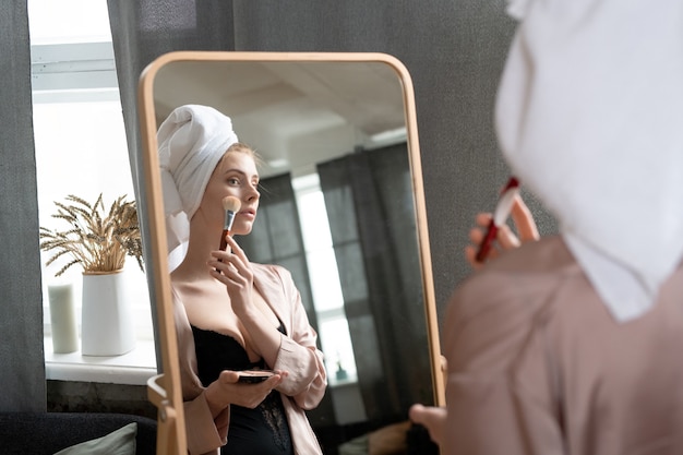 Jonge frisse vrouw in zwarte lingerie en beige pyjama die voor de spiegel in de slaapkamer staat en poeder op het gezicht aanbrengt na de ochtenddouche