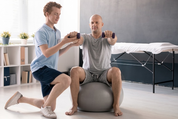 Jonge fysiotherapeut in vrijetijdskleding die mannelijke patiënt helpt om met halters te oefenen en zijn rechterarm te ondersteunen tijdens de training op fitball