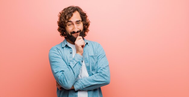 Jonge gebaarde gekke mens die gelukkig en met hand op kin glimlachen kijken, of een vraag benieuwd zijn, die opties vergelijken tegen vlakke kleur