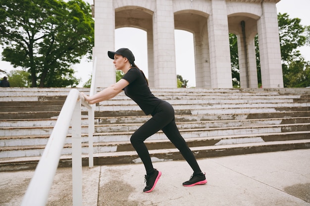 Jonge geconcentreerde atletische mooie brunette vrouw in zwart uniform en pet die sportrekoefeningen doet, opwarmen voordat ze in het stadspark buiten rennen running