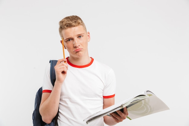 jonge gefrustreerde mannelijke student die rugzak draagt die terwijl het bestuderen met handboeken en pen in handen denkt die op wit worden geïsoleerd