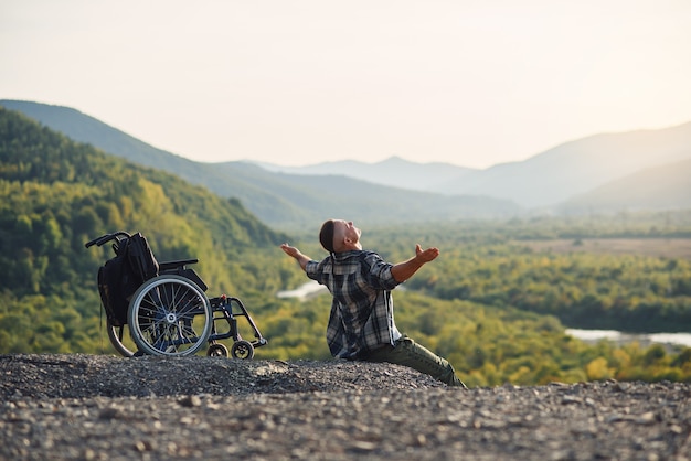 Jonge gehandicapte man zittend op de berg in de buurt van rolstoel en opgeheven handen omhoog genieten van frisse lucht en natuurschoon