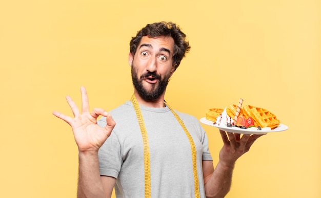 jonge gekke bebaarde man die een gelukkige uitdrukking op dieet heeft en wafels vasthoudt