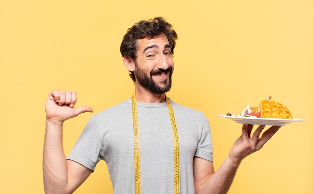 Jonge gekke bebaarde man die een gelukkige uitdrukking op dieet heeft en wafels vasthoudt