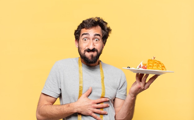 Jonge gekke bebaarde man die een gelukkige uitdrukking op dieet heeft en wafels vasthoudt