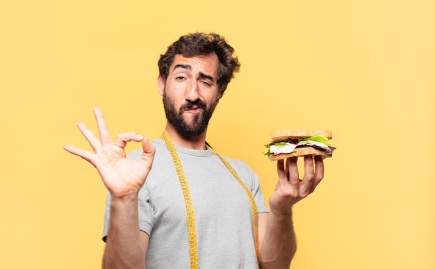 Jonge gekke bebaarde man die een gelukkige uitdrukking op dieet is en een broodje vasthoudt