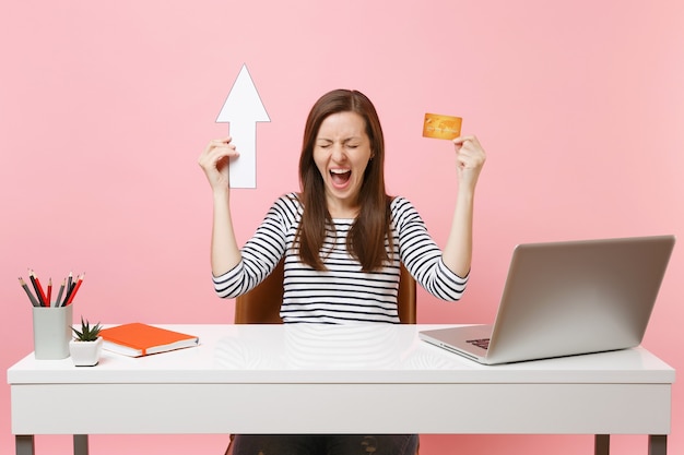 Jonge gekke winnaar vrouw schreeuwen houden pijl, creditcard zitten en werken op kantoor met hedendaagse pc-laptop geïsoleerd op pastel roze achtergrond. Prestatie zakelijke carrière concept. Ruimte kopiëren.