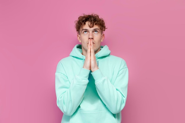 Jonge gekrulde man in mint hoodie bid en hoop op geluk op roze geïsoleerde achtergrond