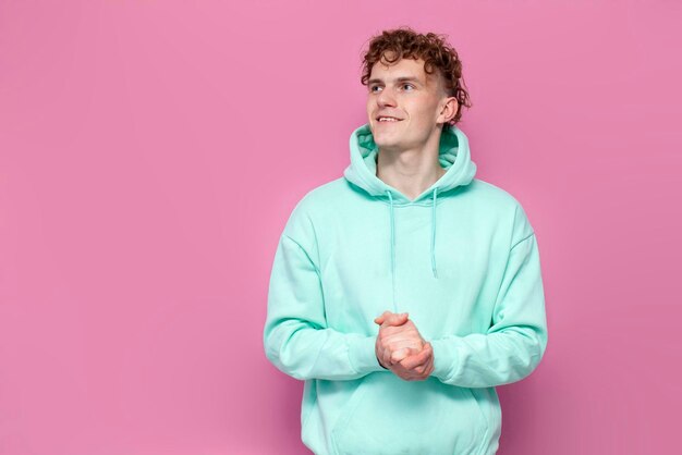 Jonge gekrulde man in mint hoodie glimlacht en kijkt weg op roze geïsoleerde achtergrond