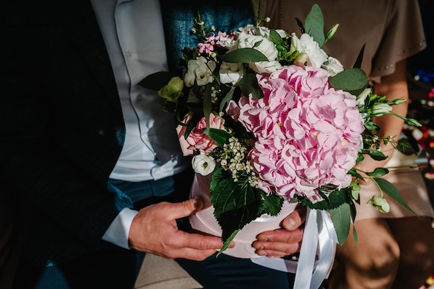 Jonge geliefden paar zitten en houden in handen stijlvol boeket van roze hortensia rozen witte anjer en groene bloemen en greens met linten Close-up bruiloft decor Artwork zijaanzicht