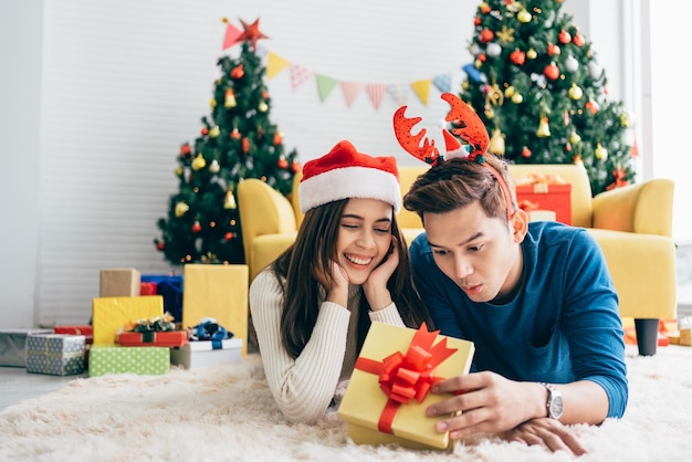 Jonge gelukkige Aziatische vrouw die een kerstmanhoed draagt met haar vriendje met een kerstcadeau terwijl ze op het tapijt ligt en naar de camera kijkt met een kerstboom op de achtergrond