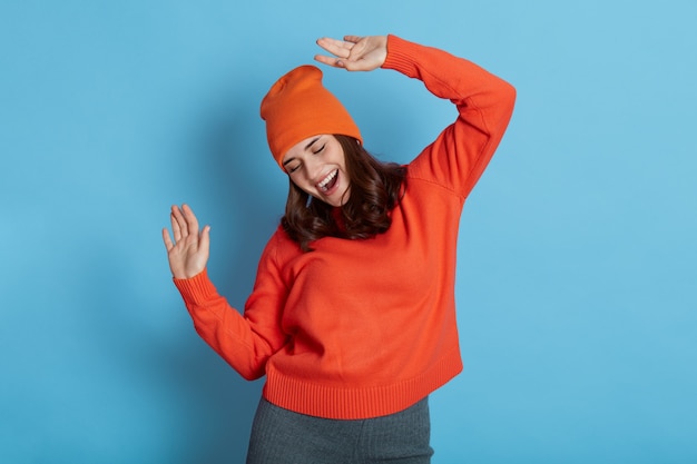 Jonge gelukkige blanke vrouw met donker haar, dansen en heeft plezier, houdt de armen omhoog, draagt trui en pet